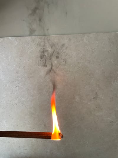 The image shows a close-up, eye-level view of an incense stick that is burning. The stick is dark brown and cylindrical. The tip of the stick is glowing orange and yellow, indicating it is aflame. A plume of grey smoke is rising from the burning tip and dissipating against a light-colored, textured surface, possibly tile or stone. The background is out of focus but appears to be a wall or surface with a marbled or speckled pattern. There is no visible indication of the location within the city of Cahul, Moldova. The scene is static and shows no people or other living beings.