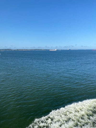 The media file depicts a wide, blue body of water with a distinct white wake trailing from the right side of the frame, indicating movement. The water exhibits ripples and texture, suggesting it is either choppy or being disturbed by a vessel. In the distance, along the left horizon, a thin strip of land is visible with what appears to be trees and some structures, suggesting a coastline. Several boats, including what seem to be larger vessels, are scattered along this distant shoreline. The sky above is a clear, bright blue, with a hint of haze or very faint clouds near the horizon.

The scene suggests a daytime activity, likely a boat trip or observation from a moving vessel, given the prominent wake. The weather appears clear and sunny. The presence of boats and the coastline hint at a maritime or coastal setting. There is no visible text in the media file.