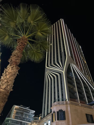 A low-angle shot at night showcases two modern buildings, one with a distinctive curved design and illuminated horizontal stripes, and the other with a more traditional facade. A palm tree, positioned in the foreground, partially obscures the base of the curved building, adding a tropical touch to the urban scene. The dark sky serves as a contrasting backdrop, enhancing the glow of the buildings' lights.