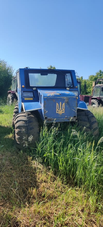 The image displays a robust, multi-wheeled blue vehicle, likely an all-terrain vehicle or specialized tractor, parked in a sunny, grassy field. The vehicle features a cabin with large windows and bright LED lights mounted on the front. Its front panel, which appears to be a perforated grille, is painted blue and prominently features a large, yellow Ukrainian national emblem (Tryzub). Faint, faded artistic designs, possibly a flower, are also visible on the side of the blue vehicle.
In the background, parts of other heavy machinery or tractors are visible, suggesting the location could be a storage yard or an area for agricultural/utility equipment. The foreground and mid-ground are dominated by tall, vibrant green grass mixed with patches of drier, brownish grass. A clear, bright blue sky with no clouds forms the upper portion of the image. The sunlight indicates it is daytime. The scene suggests a rural or semi-rural setting consistent with the provided location context of Rizunenkove, Ukraine. There are no people visible, and the vehicles appear stationary.