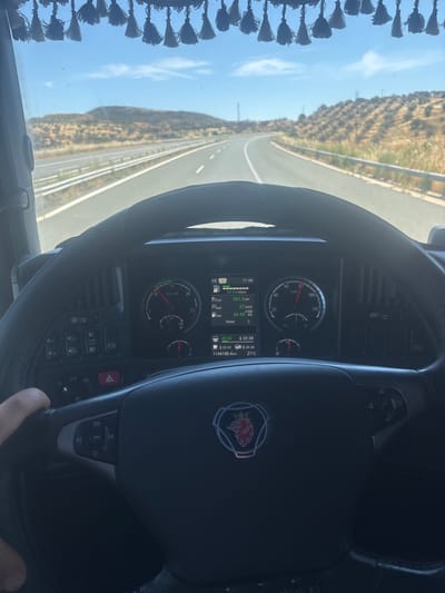The image shows a driver's perspective from inside a truck, looking out at a highway. The truck's steering wheel and dashboard are prominently featured in the foreground. The dashboard displays various gauges and a digital screen showing information such as speed (indicated around 60 km/h), trip distance (581.8 km), fuel consumption (33.5 L/100km), time (11:44), and temperature (27°C). The exterior view from the windshield reveals a sunny day with a clear blue sky, rolling hills covered in dry vegetation, and a multi-lane highway stretching into the distance. There are no people visible in the image other than the driver's hand holding the steering wheel. The truck's interior includes decorative tassels hanging from the top of the windshield. The location is identified as Alexandroupolis, Greece.