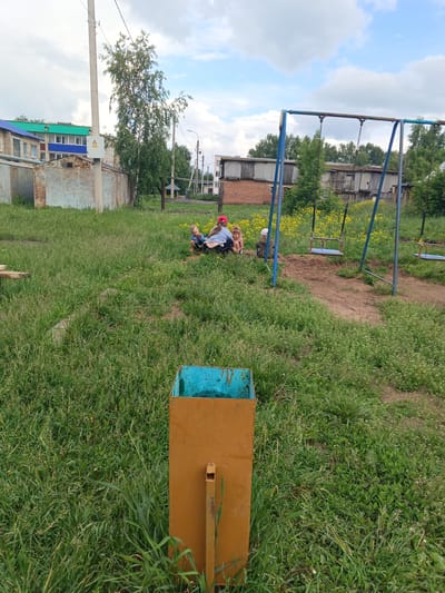 In Tolbazy, Russia, on a cloudy day, a group of four young children are gathered on a grassy area near a swing set. One child, wearing a red cap, is sitting and looking to the side. Another child, also wearing a red cap, is partially visible sitting behind the first. A third child, in a white cap, is seated facing away from the camera. The fourth child, a toddler, is seated and facing the other children. In the foreground, an orange and blue trash receptacle stands on the grass. In the background, a swing set with two empty swings is visible, along with several low-rise buildings and trees.