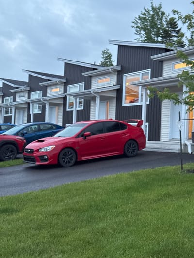 A red Subaru WRX sedan is parked on a paved driveway in front of a row of modern townhouses. The townhouses have dark grey siding and white trim, with angled roofs. The car has a large rear spoiler and black wheels. To the left of the Subaru, a blue sedan is visible. In the foreground, there is a well-maintained green lawn. The sky is overcast and grey. The scene appears to be in a residential area in Dieppe, Canada, judging by the architecture and style of the townhouses, which are common in newer developments in the region. No people are visible in the image.