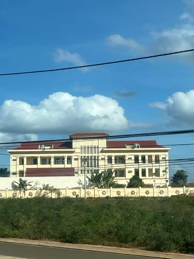 The image displays a large, multi-story building with pale yellow walls and dark red tiled roofs, situated in Trung Hồ, Vietnam, under a bright blue sky with scattered white cumulus clouds. The building features a prominent central section with vertical louvered elements and multiple wings with numerous dark-paned windows. Rooftop equipment, likely ventilation or air conditioning units, is visible on the red roofs. In the mid-ground, a light-colored perimeter wall, possibly concrete or stucco, defines the building's compound. This wall is decorated with regularly spaced circular yellow five-pointed star emblems. Tropical vegetation, including broad-leafed plants and small trees, is visible both behind the wall within the compound and as dense, overgrown green foliage in the immediate foreground. One individual is observed bent over in the overgrown area in front of the decorated wall, slightly to the left of the image center, engaged in an indiscernible ground-level activity. Dark horizontal lines, consistent with power or communication cables, traverse the upper portion of the frame, and a dark grey surface, likely a road or path, is present at the bottom edge.