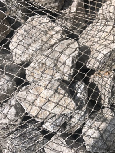 The image shows a close-up, overhead view of a gabion wall, a retaining structure filled with rocks and enclosed by wire mesh. The rocks are of varying sizes and are predominantly light gray and beige, with some darker gray ones visible. The wire mesh is a coarse, woven net with a diamond pattern, creating a textured overlay on the rocks. The lighting suggests it is daytime, casting distinct shadows from the mesh onto the rocks and creating bright highlights on the uneven surfaces of the stones. The scene appears to be outdoors, possibly a construction site or part of landscaping, but there is no specific context to determine its location within the city of Krasny, Russia. There are no people or discernible actions depicted.