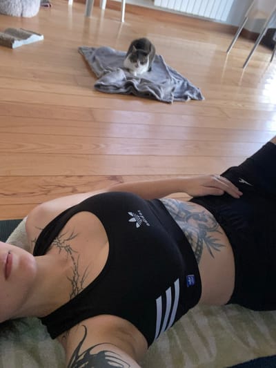 A person, viewed from an upside-down perspective, is lying on what appears to be a yoga mat on a rug. The person is wearing a black Adidas sports bra and black shorts. Tattoos are visible on the person's torso and collarbone. A gray and white cat is resting on a folded gray towel on a wooden floor. A clothing rack is visible in the background. The location is a private residence or interior setting. The specific location within the city of Villaviciosa, Spain is impossible to determine without further information or visual clues.