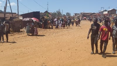 A dusty street scene features a large group of people walking in the distance. Some individuals hold umbrellas. Two men in casual attire walk towards the camera, while others stand or walk near buildings on either side of the road. The buildings appear to be modest, with some made of corrugated iron. The sky is bright and sunny. Electrical poles and wires are visible above the street.