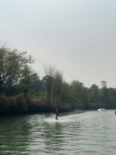 A vertical shot depicts a person water skiing in a lake, with the background featuring trees and a cloudy sky. The person is in motion, generating a spray of water. A cable system with a carrier is visible above the lake, likely used for pulling the skier. The scene is located in Tambon Nam Phrae, Thailand, as indicated by prior knowledge. A white floating object is visible in the water on the left side of the image.
