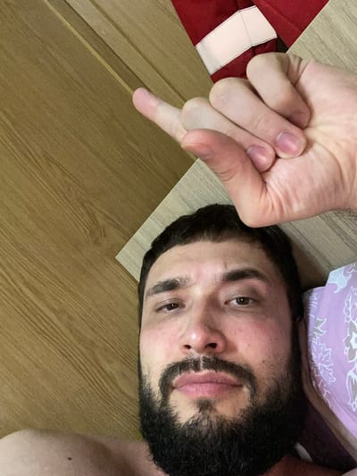 A close-up, high-angle selfie shows a man with dark hair and a beard lying in bed. He is looking up at the camera with a neutral expression, his head on a patterned pillow. His right hand is raised, with his index finger extended and pointing upwards, while his other fingers are curled into his palm. Behind him, a wooden surface is visible, with a red and white striped object at the top edge of the frame. The image has a warm, natural light.