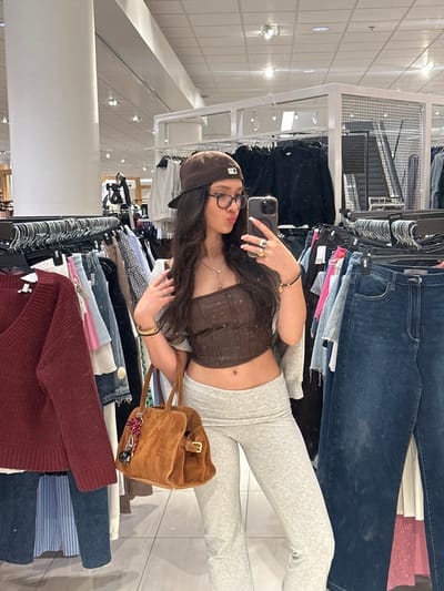 A female subject takes a mirror selfie within a retail clothing store, presumably located in Glendale, United States. She has long, dark, wavy hair, wears clear-framed glasses, and a brown baseball cap with a white stylized "NY" logo. Her attire consists of a dark brown corset-style crop top exposing her midriff, light grey high-waisted flared pants, and a light grey garment draped over her shoulders. She holds a dark smartphone in her right hand, displaying multiple rings and a dark beaded bracelet, while her left hand is positioned near her hair. Gold-colored bangles adorn her left wrist, and a delicate necklace with a small pendant is visible. She carries a light brown suede handbag featuring a buckle and a decorative charm. The background contains numerous clothing racks displaying various apparel, including a burgundy V-neck sweater and blue denim jeans. The store environment is illuminated by overhead fluorescent lighting and features white structural columns and metal grid display fixtures, indicative of a department store or large boutique.