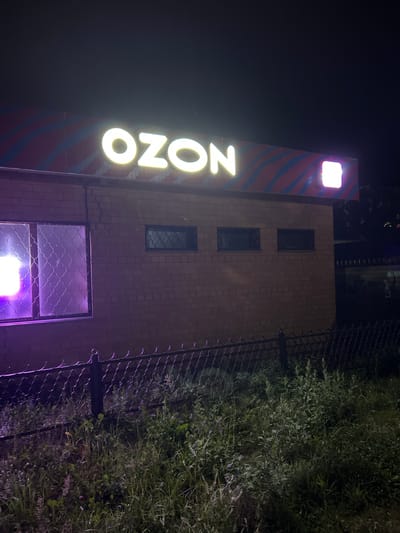 An outdoor, eye-level, night shot shows a brick building with a prominent "OZON" sign in illuminated white lettering. The sign is mounted on a horizontal band with diagonal red and blue stripes above the brick facade. To the right of the "OZON" sign, there is a small, illuminated square logo or sign in white light. The building has several windows, one large one on the left with decorative metal grates and a purple light emanating from within. Two smaller, dark windows with similar grates are visible on the right side of the building. In front of the building, there is a black metal chain-link fence, and beyond that, a dense patch of overgrown grass and vegetation. The background is dark, suggesting nighttime, with some faint lights visible in the distance to the right. The overall scene appears to be an entrance or storefront of a business in Kokhma, Russia.