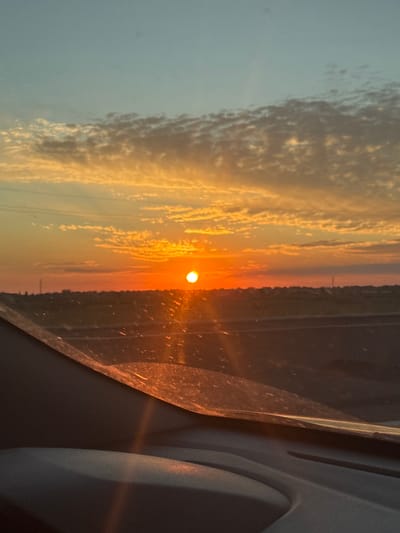 This image captures a sunset over Novotroitskoe, Russia, as viewed from inside a vehicle. The sun, a bright orange orb, is positioned near the horizon, casting a warm glow across the sky and clouds. The sky transitions from a pale blue at the top to hues of orange and pink near the horizon. Wispy clouds are scattered across the sky, illuminated by the setting sun. In the middle ground, a dark landscape with subtle details of buildings or terrain is visible, with a road or pathway running through it. The foreground is dominated by the interior of the car, with the curved shape of the dashboard and windshield reflecting the sunset's light. Small specks, possibly dust or water droplets, are visible on the windshield, catching the light. The overall scene conveys a serene and atmospheric sunset view from a moving vehicle.
