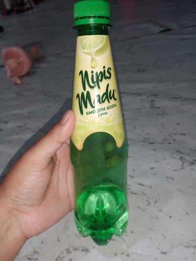 A hand holds a green plastic bottle of "Nipis Madu" smooth soda lime flavored drink. The bottle has a green cap and a label with a lime slice graphic. The background is a mottled gray surface. The image was taken in Cotseutui, Indonesia.