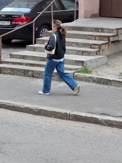The image shows a person walking outdoors next to a set of concrete stairs. The person is wearing blue jeans, a black jacket, and white sneakers. They are carrying a cream-colored bag. The setting appears to be a residential area or a public space with concrete elements. To the left of the stairs, a black car is visible. The image is taken from a slightly elevated angle, looking down at the person. The lighting suggests it is daytime, likely overcast or in shade, as there are no strong shadows. The overall scene is mundane and urban. No specific emotions are conveyed by the person's posture or expression, which is not fully visible. The text "742 AT 95" is visible on a license plate of the car. The location context is Grigorovo, Russia.