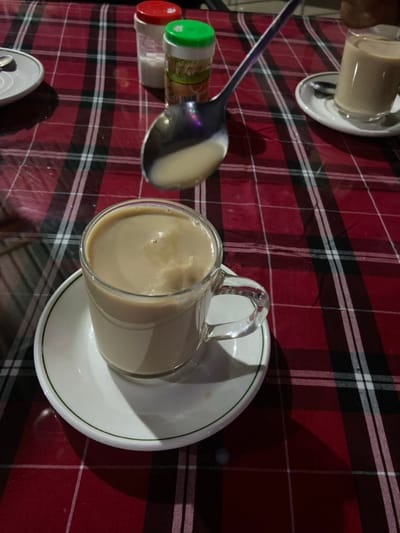 The image presents a high-angle, slightly oblique view of a table setting, likely in an indoor establishment in Bagamoyo, Tanzania. The table is covered with a vibrant red, black, and white plaid tablecloth.
The primary subject is a clear glass mug, filled with a light brown, frothy beverage that appears to be milky tea or coffee. This mug rests on a white ceramic saucer with a thin green rim. To the left of the mug, a large, reflective silver spoon lies on the tablecloth, holding a small pool of the same light brown liquid in its bowl, suggesting it has just been used for stirring or tasting the drink.
Adjacent to the spoon are two small, translucent plastic shakers or bottles, possibly containing spices or condiments. One has a bright green cap, and the other a red cap. Some faint, unreadable text or a label is visible on the green-capped shaker.
In the upper left corner of the frame, another white saucer with a similar beverage cup is partially visible, indicating multiple servings. A small portion of a silver can, possibly a soda can, can also be seen at the bottom left edge of the image.
The lighting appears subdued, casting soft shadows from the glass mug and the spoon onto the tablecloth, which suggests an evening setting or ambient indoor lighting. The scene conveys a relaxed atmosphere, perhaps a break time or a casual meal where beverages are being enjoyed. No people are visible.