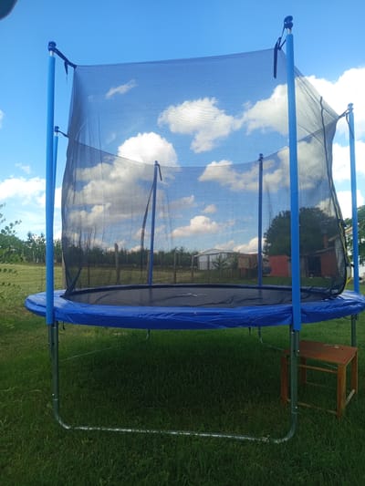 A blue trampoline with a black safety net is set up on a grassy lawn. The trampoline has blue padded poles supporting the net, and a blue padded edge covers the springs. A small, wooden stool with an orange top sits to the right of the trampoline, near one of its legs. In the background, a chain-link fence separates the yard from a more distant area with trees and some buildings, including a red-brick structure and a smaller red building. The sky is a vibrant blue with scattered white clouds, suggesting it is daytime and the weather is clear and pleasant. The scene appears to be in a rural or suburban setting, possibly a backyard. No people are visible in the image, and no text is present.