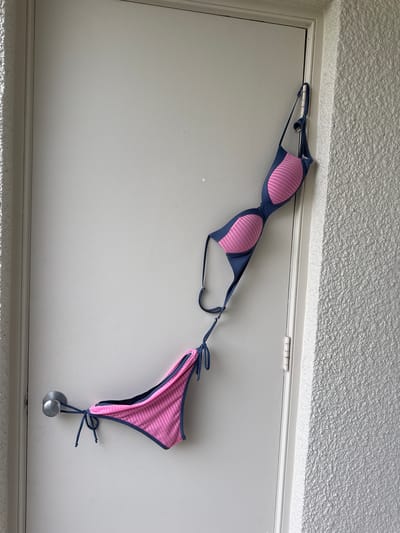 The image shows a blue and pink bikini hanging on a closed white door. The bikini top is hanging from a hook on the edge of the door, while the bikini bottom is draped over the doorknob. The door is on the exterior of a building, with a textured white wall to the right of the door. The environment appears to be outdoors, possibly a balcony or patio area. The location of this scene is the city of Boca Raton, United States.