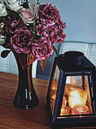 This media file captures a still life arrangement featuring a bouquet of artificial flowers and a decorative lantern. The bouquet, predominantly in shades of deep pink and burgundy with some lighter pink and white accents, is partially submerged in a glossy black vase. The flowers appear to be carnations or similar multi-petaled blooms, with some delicate greenery interspersed. The vase itself has a sleek, modern shape.
To the right of the flowers and vase is a black lantern with glass panels. Inside the lantern, several glass candle holders are visible, illuminated from within by a warm, golden light, suggesting candles are lit or the lantern is powered by an artificial light source. The candle holders have textured glass, adding visual interest.
The background consists of two distinct elements. To the left, there's a textured surface that appears to be light blue or gray, possibly a wall or upholstery with a subtle pattern. To the right and behind the lantern, a wooden surface with a prominent grain pattern is visible, suggesting a table or countertop. The overall composition is framed by the edges of these elements and the dark tones of the lantern and vase. The lighting suggests an indoor setting, possibly in the evening or a dimly lit room, given the illuminated lantern. There is no visible text in the image.