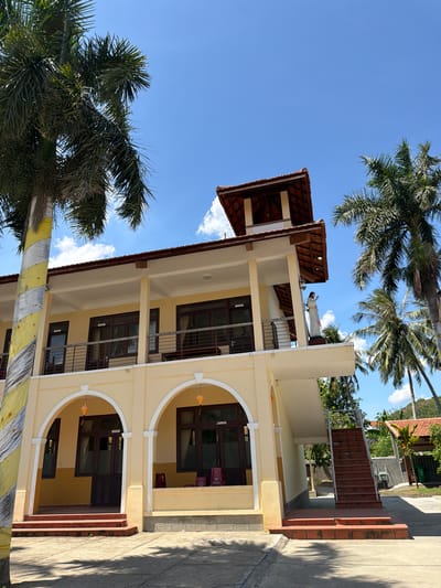 This image captures a sunny day in Hội Tín, Vietnam, featuring a multi-story building with a yellow facade and a red-tiled roof. The building has several balconies and arched entrances on the lower level, with dark-framed windows and doors. A statue of a standing figure, possibly religious, is visible on an upper balcony. Tall palm trees frame the scene, with one prominently featuring yellow and white striped decoration on its trunk. Red brick stairs lead to an external staircase on the side of the building. The ground is paved, and shadows from trees are cast across it, suggesting a tropical environment. The sky is a clear blue with scattered white clouds.