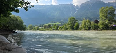 This is a landscape photograph of a wide, calm river flowing through a valley. On the left bank, a sandy shore with some debris is visible, leading to lush green trees and foliage that overhang the water. The right bank features a dense line of trees, with a small town or village nestled among them, partially obscured by the greenery. Beyond the trees, rolling green hills rise up, dotted with buildings and patches of forest. Dominating the background are majestic, towering mountains with jagged peaks, some still showing patches of snow or scree. The sky above is partly cloudy, with the sun casting a bright light that illuminates the scene. The water itself is a milky green color, with ripples reflecting the sunlight, suggesting a gentle current. Two distinct shades of water are visible in the river, with a lighter, more opaque color meeting a darker, clearer color, possibly indicating the confluence of two different water sources or sediment levels. The overall impression is one of natural beauty and tranquility, likely taken during the daytime on a clear, pleasant day. The presence of mountains and the style of architecture visible in the town suggest a location in the Alps, consistent with Innsbruck, Austria. There is no visible text in the image.