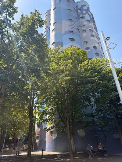 The image shows a low-angle view of the distinctive "Cité des Peupliers" apartment complex in Nanterre, France, on a clear, sunny day. The building's facade is a mosaic of blue and beige geometric patterns, with circular and oblong windows dotting its curved structure. Lush green trees with dense foliage are in the foreground, partially obscuring the building and casting dappled shadows on the paved walkway.
On the right side of the frame, a tall white pole stands, topped with a surveillance camera and a solar panel array. In the middle ground, two men are seated on a park bench beneath the shade of a tree, facing the building. To the left, a man in a light-colored t-shirt and white shorts walks along the pathway, his back to the camera. In the background, construction fencing is visible. The overall scene depicts an urban environment with a blend of modern architecture, natural elements, and pedestrian activity.