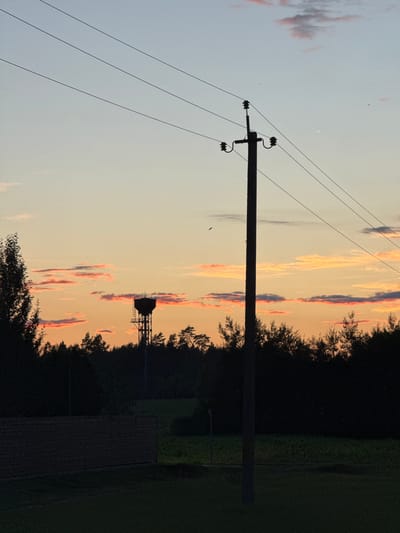 The photograph captures a sunset scene in the Neman Volost, Belarus. In the foreground, a silhouetted wooden fence runs horizontally across the lower portion of the frame. To the right of the fence, a grassy area leads towards a dark line of trees. A prominent, dark silhouetted utility pole stands to the right of center, supporting several power lines that stretch across the sky. In the mid-ground, a dense line of trees forms a dark silhouette against the colorful sky. Further in the distance, a water tower is visible as a dark silhouette against the horizon, with a structure of metal supports beneath its tank. The sky exhibits a gradient of colors, transitioning from a pale blue at the top to soft oranges and pinks near the horizon, with scattered, wispy clouds tinged with pink. The overall atmosphere is tranquil and serene, characteristic of a rural twilight.