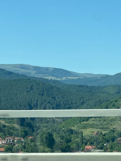 This is a landscape photograph taken from a high vantage point, possibly from a window or balcony, looking out over a lush, green mountainside. The image is oriented vertically.
On the left side of the frame, a vast expanse of clear, pale blue sky dominates. The right side of the image reveals a steep, verdant slope covered in dense foliage and scattered houses with red-tiled roofs. The houses appear nestled into the hillside, suggesting a rural or semi-rural setting. The trees are a deep, rich green, indicating a healthy and well-established natural environment. Further into the distance, successive mountain ridges recede, becoming lighter in color and less defined, hinting at hazy, atmospheric conditions or a great distance.
The scene suggests a peaceful, possibly sunny day. The quality of the light, with clear shadows and bright colors, implies it is daytime, likely mid-morning or afternoon. The presence of houses indicates a human settlement within this natural landscape. The overall impression is one of natural beauty and tranquility, with a sense of elevation and expansive views. No people or specific objects are clearly discernible. There is no visible text in the image.