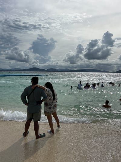 In a scenic Thai beach setting, several people are enjoying the water and shore. The day appears bright with sunlight reflecting off the water. Dark, dramatic clouds dominate the sky, suggesting a dynamic weather system. In the foreground, a couple stands on the sandy beach, with the man embracing the woman from behind. They are both looking out towards the ocean. The woman wears a floral-patterned dress, and the man is dressed in a green shirt and khaki shorts. The sea is calm with gentle waves lapping at the shore. Further out in the water, a group of people are wading and swimming, their figures silhouetted against the bright sunlight. The image captures a moment of leisure and relaxation at a tropical beach.