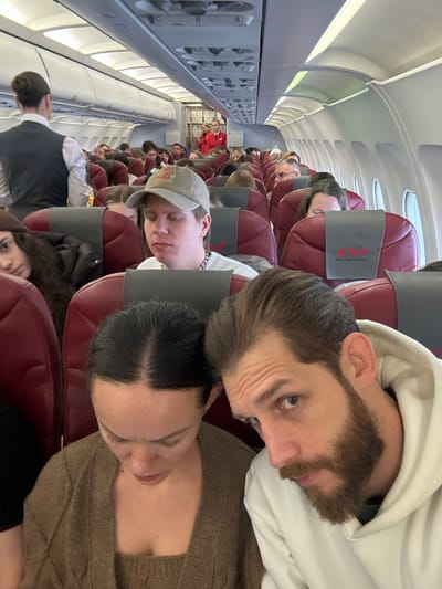 The scene depicts the interior of a commercial aircraft cabin, identified as a Nordwind Airlines flight by visible seat branding. The specified location for this event is Novogorelovo, Russia. The image captures a dense arrangement of passengers seated in dark red/maroon economy class seats with grey headrest covers, extending towards the front of the aircraft.

In the immediate foreground, a male with a brown beard and light eyes, wearing a white hoodie, is positioned on the right, looking directly towards the camera. To his left, a female with dark hair, wearing a brown knitted top, is looking downwards. Behind them, another female with long dark hair is partially visible, glancing towards the camera from the seat to the left. Central to the mid-ground, a male wearing a light-colored baseball cap with a "JESUS" graphic and a silver chain is seated with his eyes closed.

Further down the aisle on the left, a female flight attendant with her hair in a bun, wearing a dark vest over a white shirt, stands with her back mostly to the camera. In the distant background, near the front galley area, two additional female flight attendants in red uniforms are visible, one gesturing. Overhead bins and passenger service units are visible throughout the cabin. Windows on the right side of the aircraft reveal bright, ambient daylight. The cabin is filled with passengers, occupying most visible seats.