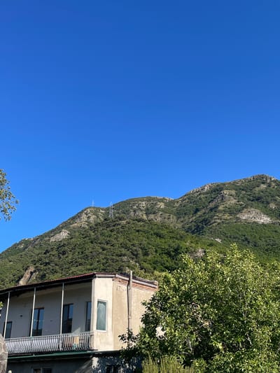 This is a landscape photograph taken on a clear, sunny day, featuring a vibrant blue sky dominating the upper portion of the frame. Below the sky, a lush green mountainside fills the middle and lower portions of the image. The mountain is covered in dense foliage and shows rocky outcrops. In the foreground, a two-story building with a cream-colored facade is visible. The building has a covered balcony on the upper floor with a white railing and several windows. Trees with green leaves partially obscure the building on the right side of the frame. Two tall, metal electrical pylons are situated on the crest of the mountain in the distance. The scene appears to be a rural or semi-rural area, possibly on the outskirts of a city. Based on the typical landscape and architecture, this image could be located in Mtskheta, Georgia, a city known for its mountainous surroundings and historical sites.