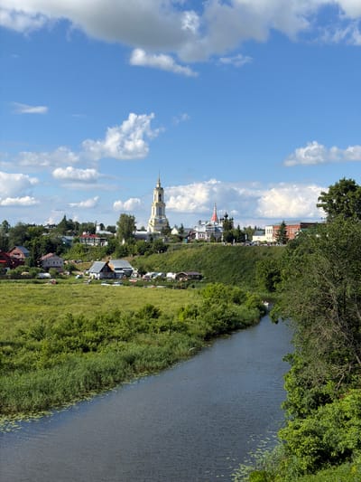 The image shows a panoramic view of the city of Suzdal, Russia, on a sunny day with scattered clouds. The scene is dominated by a river flowing in the foreground, bordered by lush green vegetation and reeds. In the middle ground, several houses and buildings are scattered across a grassy hill. A prominent yellow bell tower with a spire stands tall in the background, along with other church domes and buildings characteristic of Russian architecture. The overall impression is one of a peaceful, historic town nestled within a natural landscape.