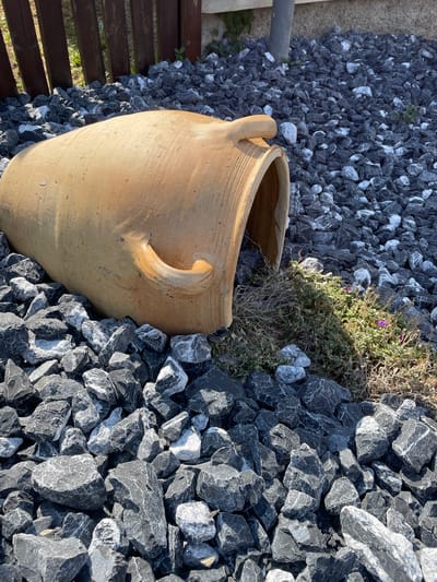 This media file is a still image taken outdoors during the daytime, likely in a garden setting. The scene is dominated by dark grey and white gravel, with a large, light brown clay pot positioned prominently in the lower left portion of the frame. The pot has two handles and appears aged, with visible cracks and texture. To the left of the pot, a section of a wooden deck or fence is visible, constructed from dark brown planks. In the upper middle of the frame, a small patch of dry-looking green and brown vegetation with tiny purple flowers is growing amongst the gravel. The sunlight casts shadows, indicating a sunny day. The overall impression is one of a decorative outdoor space, possibly a courtyard or garden path. There is no visible text in the image. The location context provided is Gerstheim, France.