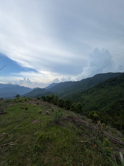 The image shows a mountainous landscape near Huế, Vietnam. A steep, green hillside slopes down to the right, covered in grass and some vegetation. A denser, darker green forest covers a ridge rising upward on the left. In the background, layers of hazy blue mountains recede into the distance. The sky is cloudy with patches of blue. The picture is slightly tilted to the left.