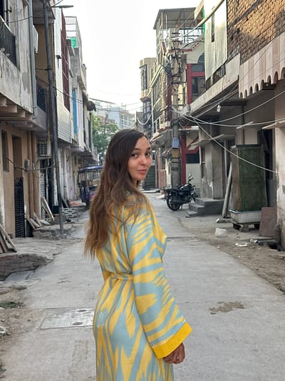 A young woman stands in a narrow street in Mathura, India. She is positioned to the right of the frame, wearing a light-blue and yellow-striped garment with yellow cuffs. Her long brown hair flows down her shoulder. The street, made of concrete, is flanked by aged buildings. The buildings on both sides are a mix of residential and commercial structures, with visible signs of wear and tear. Utility wires crisscross overhead, adding to the cluttered urban atmosphere. A parked motorcycle can be seen further down the street. The architecture features a mix of brick and concrete, with windows and balconies lining the facades. The overall environment suggests an older, densely populated urban area.