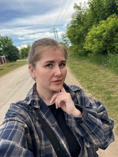 The media file is a portrait-oriented selfie of a young woman outdoors. She is wearing a blue and tan plaid flannel shirt over a black top, with a black strap visible across her chest that appears to be part of a bag. Her blonde hair is pulled back, and she has on light makeup, including mascara and lipstick. She is touching her chin with her right hand. The background shows a path or dirt road bordered by grass and trees, with a cloudy sky visible above. Power lines are also present in the sky. The lighting suggests it is daytime, possibly late afternoon. The setting appears to be rural or semi-rural. Visible text on the strap reads "DAVID JONES". The overall mood seems casual and relaxed.