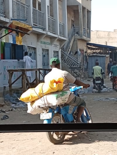 This image depicts a street scene in Kofar Ruwa, Nigeria, under bright daylight. In the immediate foreground, a dark horizontal bar partially obstructs the view. A light blue motorcycle, viewed from its rear, occupies the center-foreground. It carries a substantial yellow sack strapped horizontally onto its rear rack, with a white patterned sack secured underneath. The motorcycle's license plate, partially visible, reads "JIGAWA GND 338GA." The male rider, clad in a light-colored shirt and a green baseball cap, is seated on the motorcycle, his bare feet visible near the footrests.
To the left of the scene, a person in a long white garment is positioned beside a stack of large, flat panels, some labeled "TINONG ALUMINUM COMPOSITE PANEL." These panels are arranged near a simple wooden frame structure. Behind this activity, a multi-story concrete building with multiple ground-floor entrances and upper-level balconies with metal railings is visible. A sign on this building reads "SRI UMARA ENTERPRISES."
Further into the background and to the right, another multi-story concrete building features an external metal staircase leading to upper floors, with various items, possibly clothing, draped over its railings. The unpaved ground throughout the scene appears dusty or sandy. Several other individuals and motorcycles are present further down the street. One man in a lime green shirt is riding a dark motorcycle, also seen from the rear, while another man in a blue or green shirt is visible standing or walking. Simple structures, possibly market stalls with corrugated metal roofs, are discernible in the right background.