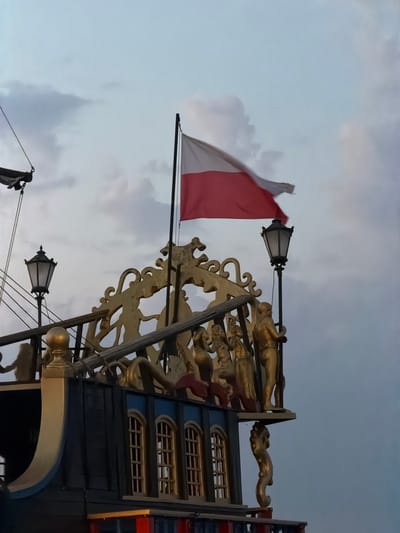 The image shows the top of a decorative ship structure, likely a replica or themed attraction, against a cloudy sky. A Polish flag, horizontally divided into white and red, is flying from a flagpole. The ship's upper deck features ornate golden decorative elements, including figures that appear to be statues of people. A black lamppost with a lantern is also visible on the deck. The ship's structure is dark blue with gold trim and arched windows. This structure is located on the Sopot pier (Molo w Sopocie) in Sopot, Poland, a popular tourist destination. The lighting suggests it is either early morning or late evening, with soft, diffused light from the overcast sky.