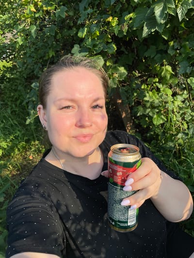 A medium shot taken from a low angle shows a woman holding a can of drink in her right hand. She is wearing a black t-shirt with small white dots and a silver chain necklace. Her hair is pulled back, and she has light pink nail polish on her fingernails. The background consists of green foliage, possibly from a garden or park. The lighting suggests it is daytime, with sunlight illuminating the scene. The location is identified as Baldeyka, Russia.