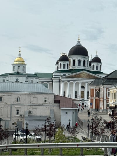 The image depicts a view of Arzamas, Russia, showcasing architectural landmarks. A prominent church structure with multiple domes, some black and some gold, stands out against a cloudy sky. Adjacent buildings feature architectural details such as columns and decorative facades. The cityscape includes streets with pedestrian traffic, streetlights, trees, and parked cars. A metallic railing runs along a green area on the side.