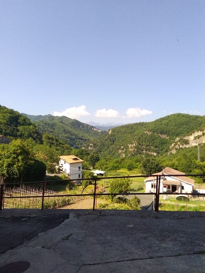 The image captures a scenic view of the mountainous landscape surrounding Valle Castellana, Italy. In the foreground, a concrete area transitions into a metal railing, behind which a small cultivated garden is visible. Further back, nestled among lush green trees, are two white houses with terracotta roofs. The houses appear to be residential dwellings. The background is dominated by rolling green hills and mountains, with a clear blue sky above, dotted with a few wispy clouds. The overall impression is one of rural tranquility and natural beauty. There are no people visible in the image.
