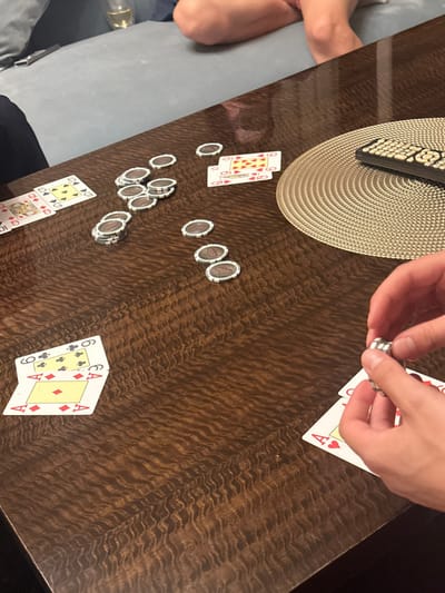 The image displays an indoor scene in Vandœuvres, Switzerland, focusing on a polished dark brown wooden table with a distinctive rippled grain pattern. A card game is underway. Scattered across the table are numerous circular, silver-grey game tokens, some in small stacks, others individually. Playing cards are also visible: a partial fanned hand showing a 5 of Spades and a 9 of Spades in the upper left; two cards, a 6 of Hearts and a 9 of Hearts, in the mid-right; and two overlapping cards, an Ace of Diamonds and a 6 of Spades, in the lower right. On the right side of the table, a light-colored, circular woven placemat holds a black remote control with light-colored buttons. In the lower right foreground, a pair of human hands is actively holding and manipulating a stack of the silver-grey game tokens. The background, blurred, reveals a light grey upholstered sofa with the lower torsos and bare legs of at least two individuals resting upon it. A glass containing a clear, light-colored beverage is partially visible on the table near the sofa.