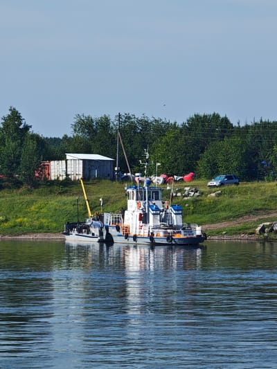 The image depicts a sunny day scene on a body of water, likely a river, with a tugboat and a smaller vessel docked on the left. The tugboat, white with blue accents and a gray hull, displays Russian text on its stern that reads "АРКАДИЯ ПАЛЕЛИКОВ". It has several decks, railings, and equipment, including a crane. The smaller vessel, to the left of the tugboat, is white with black trim and has a crane-like apparatus extending from its bow. Both boats are moored close to the shore, which is lined with green grass and trees. In the background, several shipping containers, one red and two white, are visible on the grassy embankment, along with a small white building with a corrugated roof. Further back, a dense line of green trees forms the horizon. A silver car is parked on the grassy slope to the right, behind the tugboat. Several red buoys are scattered on the embankment near the car. The water is calm with visible ripples and reflections of the sky and the vessels. The sky is clear and bright blue. The scene appears to be in or near the village of Galanino, Russia, judging by the context provided.