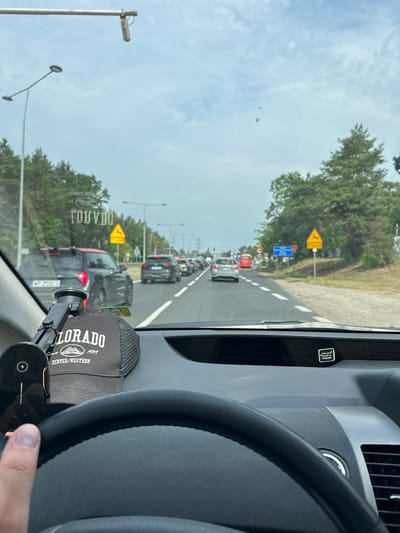 The image is taken from the driver's perspective inside a car, looking forward at a road with traffic. The driver's steering wheel and part of the dashboard are visible in the foreground. A phone mount with a wireless charger is attached to the dashboard. A black baseball cap with "COLORADO DENVER/WESTERN SINCE 1989" printed on it is resting on the dashboard. The car is on a road with multiple lanes and a line of cars ahead. There are trees and a cloudy sky in the background. On the right side of the road, there are traffic signs. One sign is a yellow triangle with a deer symbol, indicating a potential hazard. Another sign is a blue rectangular sign. The environment appears to be semi-urban or suburban, with trees lining the road. A text reflected on the windshield of the car reads "TOHYDO". This scene likely captures a moment of traffic congestion on a road in or near Łomianki, Poland, possibly on a highway or a major route due to the multi-lane road and traffic signs.