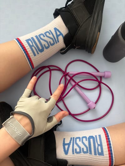 A top-down shot depicts a pair of legs wearing white socks with "RUSSIA" printed in blue and a red and blue stripe at the top, along with a pair of black athletic shoes. Next to the legs is a purple jump rope, a grey water bottle, and a hand wearing a grey exercise glove. The background is a light blue surface with a subtle patterned texture. The location cannot be determined based on the objects in the scene. Therefore, the location of Анкудиновка, Russia, cannot be confirmed.