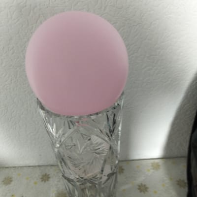 A pink ball rests on the rim of a clear, ornate crystal vase. The vase appears to be empty and is positioned on a surface with a patterned tablecloth. Behind the vase and ball, a textured white wall serves as the backdrop. The scene is lit with a soft light, creating some shadows and highlights on the objects and the wall. There are no people present in the image. The location is not identifiable from the image.