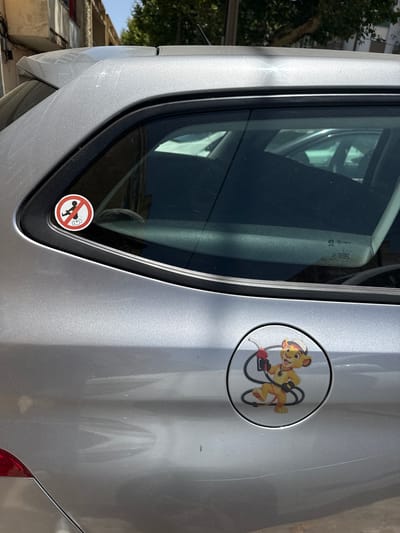 The image shows a silver car parked on a street in Almada, Portugal, under a clear sky. On the car's rear passenger window, a circular sticker with a red border depicts a figure in a squatting position with lines indicating a release of gas or air, accompanied by a red circle with a diagonal line through it, signifying a prohibition. Below this sticker, on the fuel cap, is another circular sticker featuring a cartoon lion character, possibly Simba from The Lion King, wearing a white hat and holding a red fuel nozzle connected by a black hose. The lion is yellow with orange stripes and a light brown muzzle, and appears to be in motion, with one leg raised. The surrounding environment consists of a building with a red facade visible in the background and trees. The car itself appears to be a station wagon, with its rear light visible in the lower left corner.