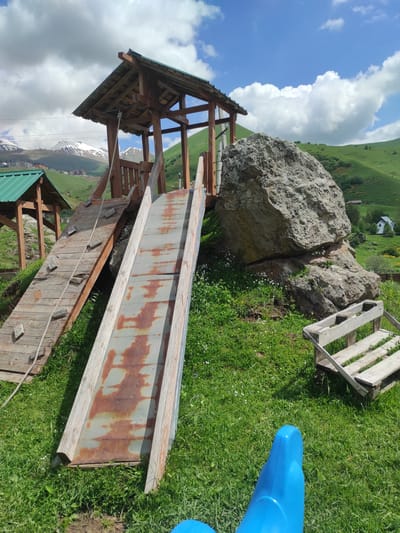 This image depicts an outdoor playground structure set against a backdrop of rolling green hills and snow-capped mountains, suggesting a location in a mountainous region such as Kumlistsikhe, Georgia. The playground features a wooden fort with a green roof, a rusted metal slide, and a wooden climbing ramp with rope. A large boulder is situated next to the fort, and a simple wooden bench made from pallets rests on the grass nearby. The foreground is occupied by a portion of a blue plastic ride-on toy. The sky is partly cloudy with patches of blue. No people are visible in the image. The overall scene conveys a rustic and somewhat weathered appearance of a playground in a natural, rural setting.