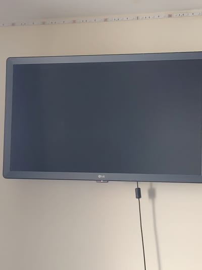 A television screen displays a blank, dark gray image. The television is mounted on a pale beige wall. Along the top edge of the wall, a strip of small, illuminated LED lights is visible. The bottom right corner of the TV screen shows the LG logo. A black cable hangs down from the bottom of the TV, casting a long shadow on the wall. The scene appears to be indoors, in a room that is not brightly lit. No people or actions are depicted. The specific location within Northampton, United Kingdom, cannot be determined from the visual information provided.