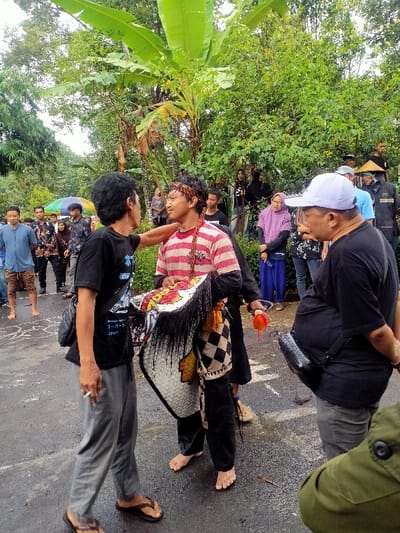 An outdoor scene in Kedawung, Indonesia, depicts a gathering on a wet, dark paved surface, bordered by dense green foliage, including prominent banana trees. In the foreground, three adult males are positioned. On the left, a male with dark curly hair, wearing a black t-shirt, grey sweatpants, and black flip-flops, holds a lit cigarette in his right hand. His left hand rests on the right shoulder of the central male. The central male, also with dark hair and a patterned headband, wears a red and white striped t-shirt and dark pants. He is barefoot. He holds a white and black woven 'kuda lumping' (flat horse puppet) costume element against his left side and a small orange and red feathered object in his right hand. A beaded necklace is visible around his neck. To the right, partially visible, a third male wears a light-colored baseball cap, a black t-shirt, and grey pants, carrying a black shoulder bag. He faces away from the camera, observing the central interaction. In the midground, a diverse crowd of onlookers is visible. To the left, a group of men, some barefoot, stand near a multi-colored umbrella. To the right, individuals, including women wearing hijabs, are positioned against the green backdrop. White chalk-like markings are present on the wet pavement in the mid-left.