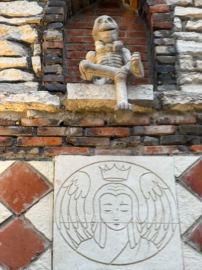 The image depicts architectural details of a building in Domingo Arenas, Mexico. On the left, a stone sculpture of a skeleton holding an object in its hand is embedded in a wall composed of bricks and irregularly shaped stones. To the right, a square stone carving features a circular design of an abstract human figure with wings, surrounded by diamond-shaped terracotta tiles.